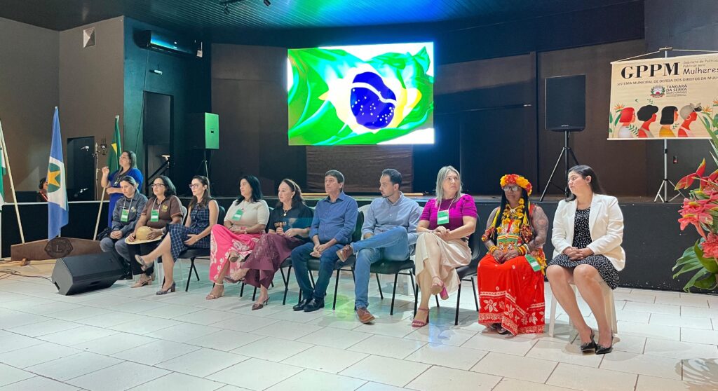 Autoridades e representantes de diferentes segmentos participam do evento para discutir políticas públicas voltadas às mulheres.
#Paratodosverem: Em uma fileira, onze pessoas estão sentadas lado a lado Entre elas, uma mulher indígena com vestes tradicionais e uma intérprete de libras em pé ao fundo.
