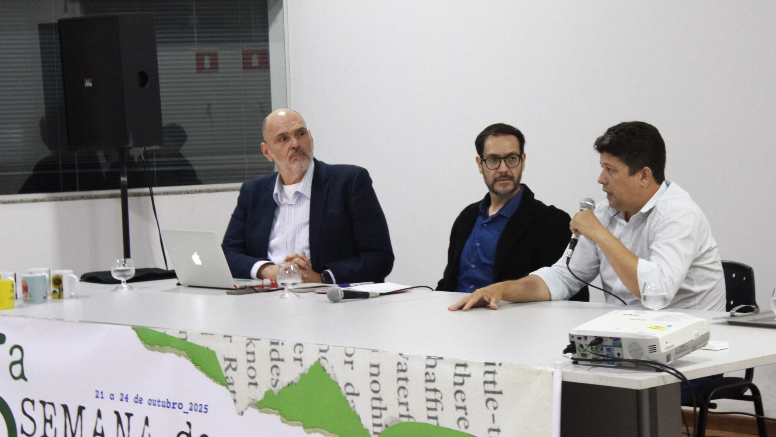 À esquerda, o professor Carlos Filho; ao centro, o professor Ulisflávio Evangelista; à direita, Adriano Soares. Os três estão sentados, e Adriano segura um microfone sendo observado pelos outros dois professores.