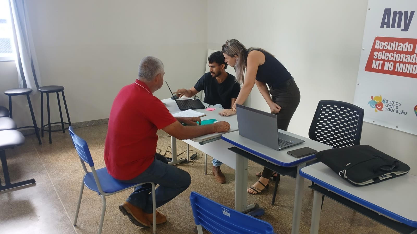 Dois servidores públicos sendo um homem e uma mulher, vestidos de preto realizando o cadastro de um senhor, produtor rural vestido de camisa vermelha e calça jeans azul. O espaço utilizado é uma sala de aula com algumas mesas espalhadas em tons de azul e cinza.