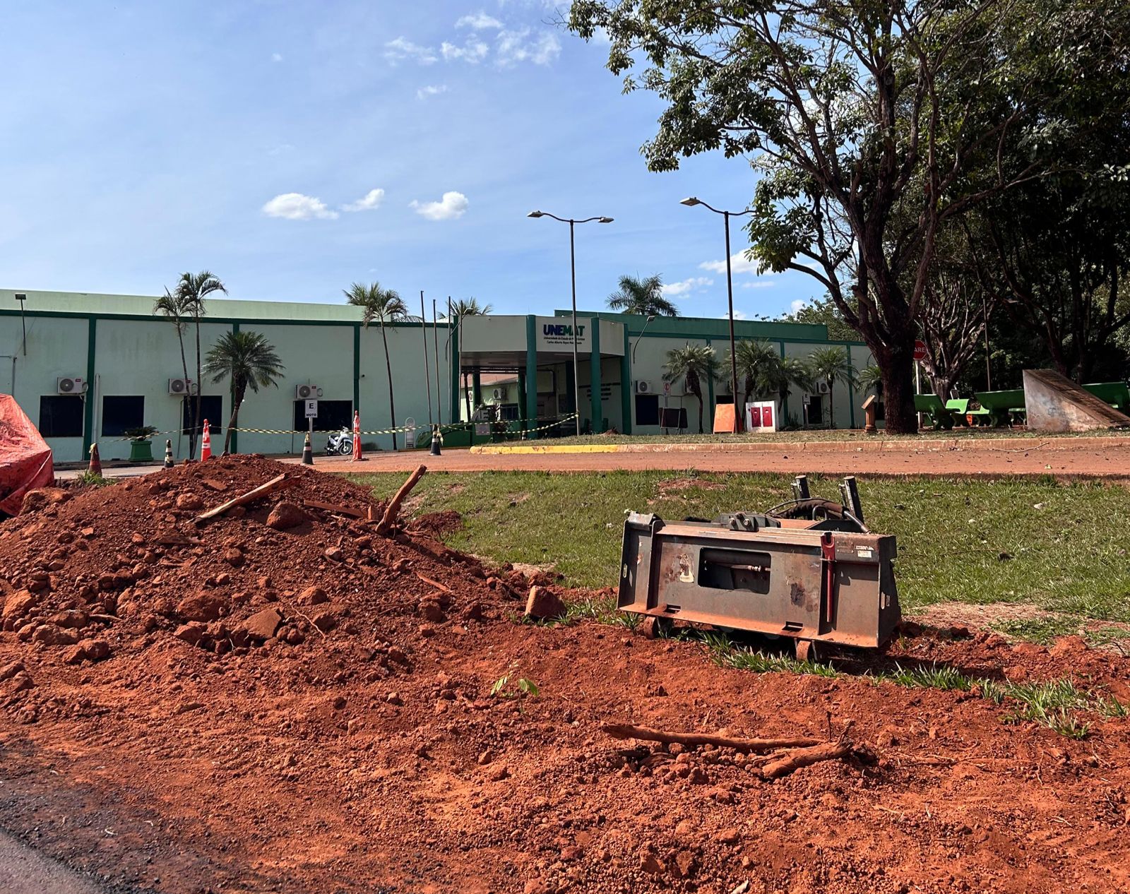 Foto da obra no campus da Unemat de Tangará da Serra. A imagem contém muita terra na frente do campus, contém cones e faxas que sinalizam a obra no geral. Atrás, está o campus da Universidade do Estado de Mato Grosso, identificado com tons de verde claro e escuro.