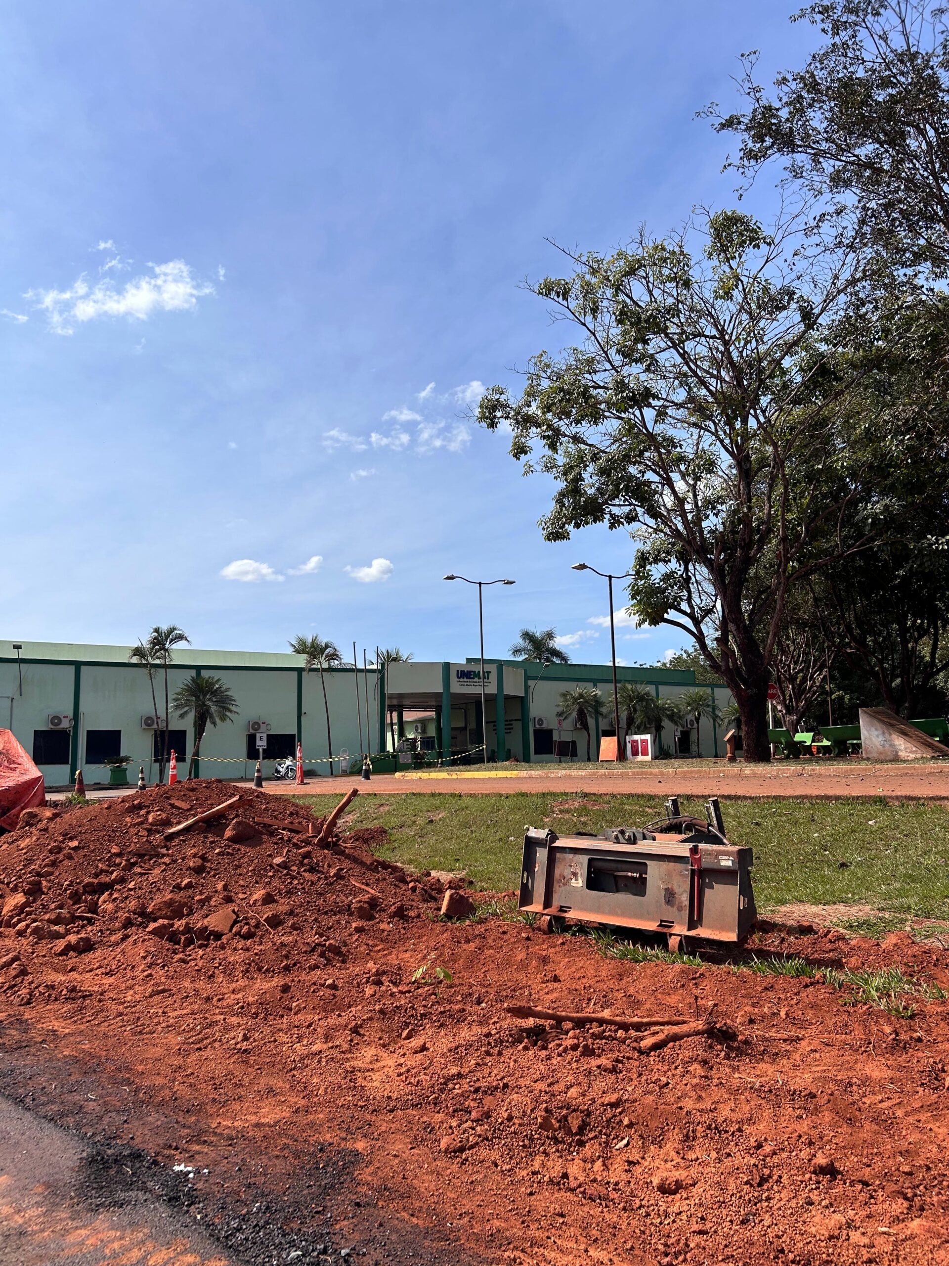 Imagem mostra campus de Tangará da Serra Unemat ao fundo, dando destque as construções que estão sendo realizadas na universidade. A frente da imagem apresenta morros de terra, fitas e cones de sinalização, com o dia ensolarado e ceú sem muitas nuvens e árvores no canto direito.