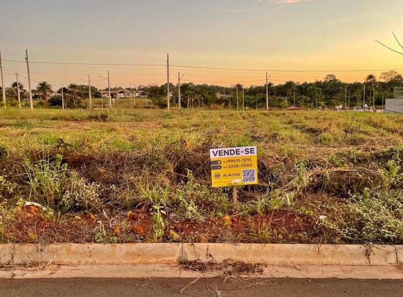 A imagem apresenta uma placa de comercialização de lote. O céu está um misto de cores que variam entre azul claro e laranja. A placa está fixada no chão com matos em sua volta