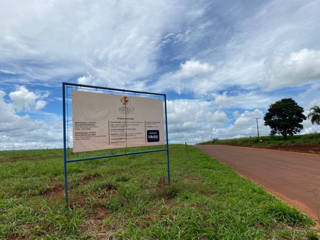 Na foto temos a imagem de uma placa localizada em um terreno com mato verde e ao lado uma estrada de terra vermelha, o céu está bem azul e ensolarado. A placa está falando sobre o local ser um loteamento denominado “Reserva do parque II”, que está aberto para vendas.