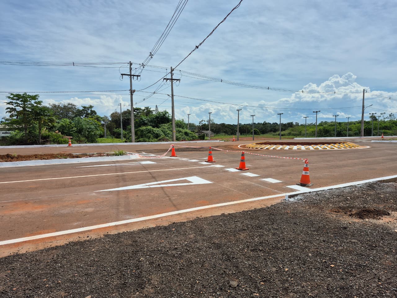 Ao centro da imagem, a via asfaltada possui sinalização horizontal branca recém-pintada, incluindo faixas de orientação e marcações de tráfego. Cones laranja e fita de isolamento delimitam um trecho ainda em obras. À direita, há uma rotatória circular com sinalização amarela e branca nas bordas. Ao fundo, diversos postes de iluminação e redes elétricas acompanham a extensão da via. A paisagem compõe a imagem com uma vegetação verde com árvores.