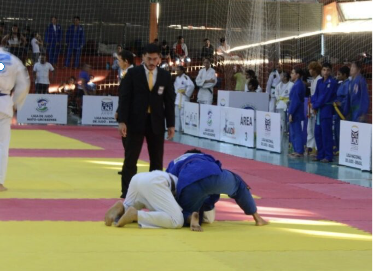 Na foto aparecem dois atletas de judô, um com kimono azul e outro, Enzo Gomes, de kimono branco. Um árbitro de terno acompanha disputa, enquanto outros competidores aguardam ao redor. 