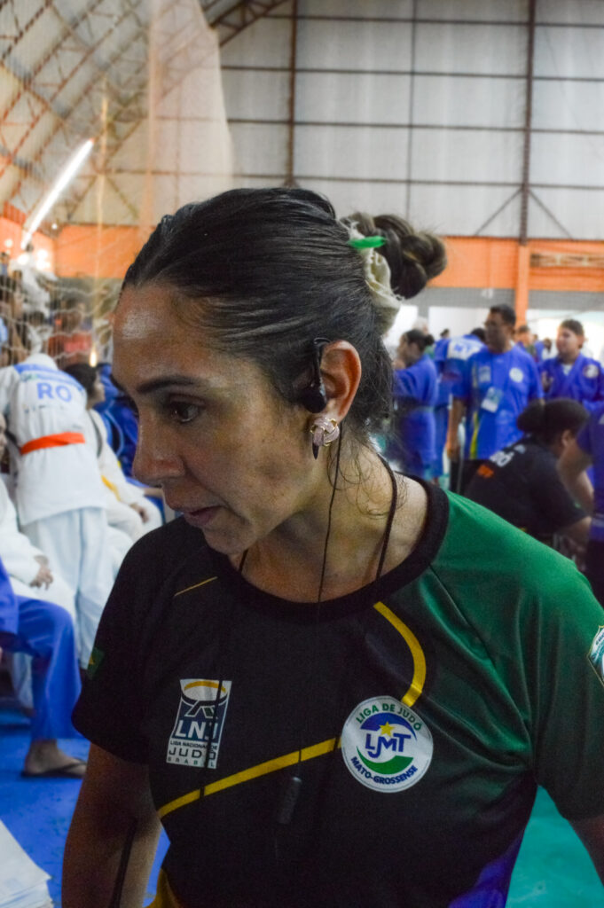 Na foto aparece uma das pessoas responsáveis pela organização do evento esportivo. . Ela Vesta uma camiseta com as cores verde e preta, usa um fone de comunicação que a auxilia no acompanhamento e coordenação do evento. Ao fundo, atletas de judô em kimonos azuis e brancos.