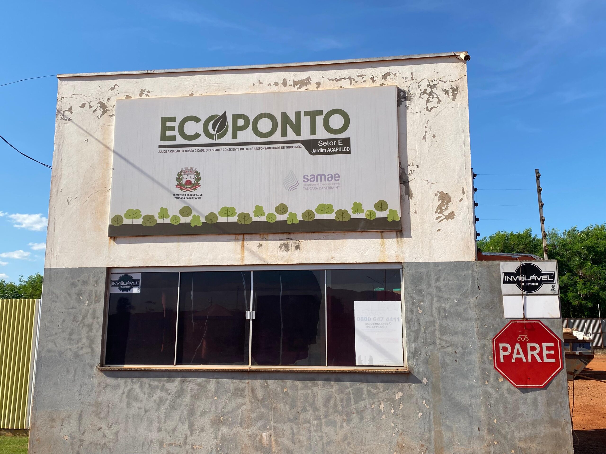 Na imagem temos a faixada do Ecoponto do Setor E, localizada no jardim Acapulco, a mesma é uma pequena guarita onde o fiscal fica. De fundo temos um céu azul e alguns galhos de árvores. Na faixada temos o nome "ECOPONTO" com grafias que remetem ao ecológico, mais abaixo temos uma ampla janela e uma placa de pare vermelha.