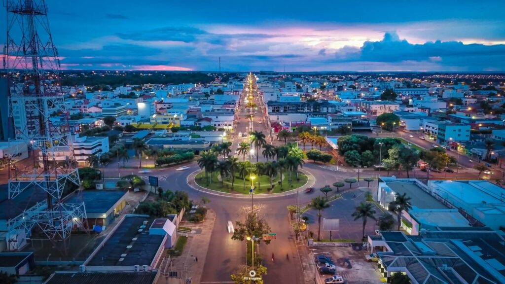 A foto mostra uma vista aérea de Tangará da Serra. No centro tem rotatória pricipal da avenida Brasil, com palmeiras e postes de luz. 