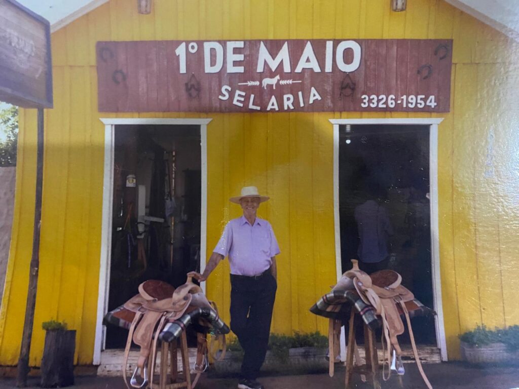 Na imagem aparece o senhor Tamir Torres em frente a Selaria 1º de Maio, ao lado dele duas selas para cavalo. A selaria é uma casa simples de madeira, pintada de amarelo.